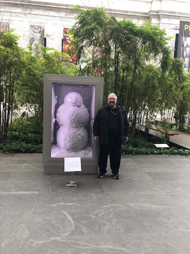 Jeffrey F. S. Neumann standing beside the glass-encased Mr. Snowmann sculpture in a London atrium exhibition, 2015