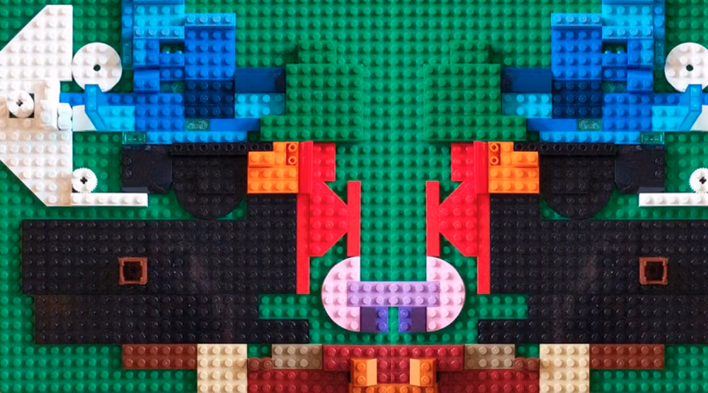A symmetrical face built on a green Lego baseplate, in blue, red, black and pink bricks.